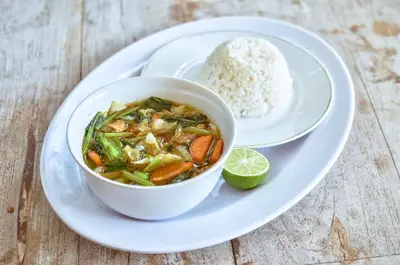 a bowl of soup on a plate with rice and a lime at Palm Garden Bali in Nusa Dua