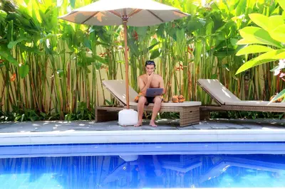 a man sitting in a chair with a laptop next to a swimming pool at Kaniya bali in Nusa Dua