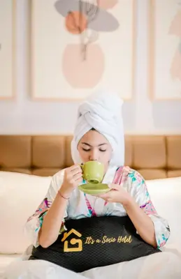 a woman sitting on a bed drinking from a green cup at Graha Socio Hotel Nusa Dua Bali in Nusa Dua