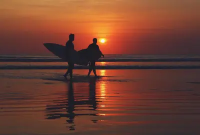 two people walking on the beach at sunset with their surfboards at KUTA - 4BR Villa with Private XL Pool in Kuta